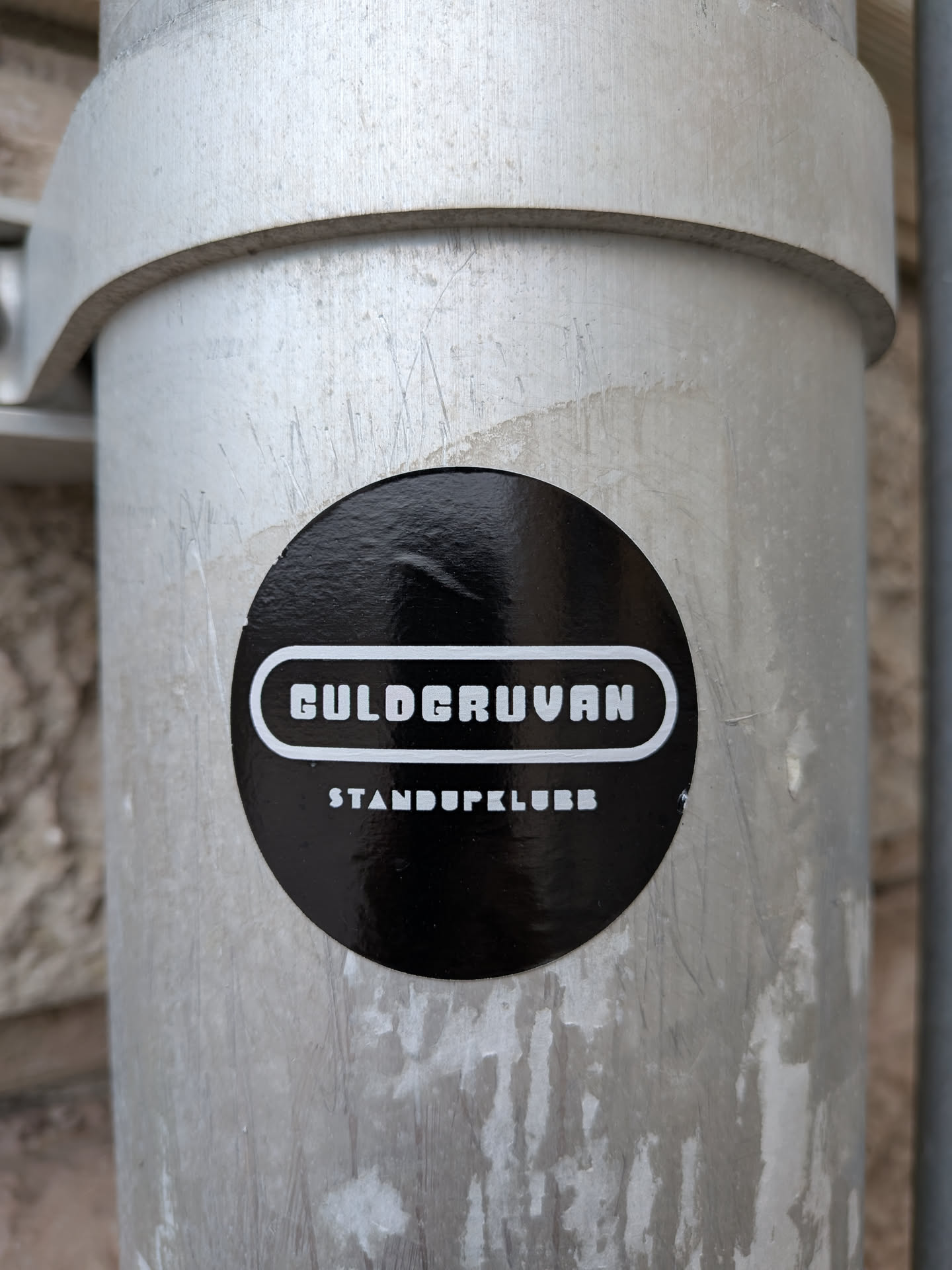 The sticker is circular and predominantly black with white text. The text is arranged in two lines. The top line features the word 'GULDCURVAN' in a stylized, bold, sans-serif font with a white outline. Below this, in smaller, all-caps sans-serif font, is 'STANDUPKLUBB'. The sticker has a glossy finish and is slightly curved, adhering to a metallic cylindrical surface.