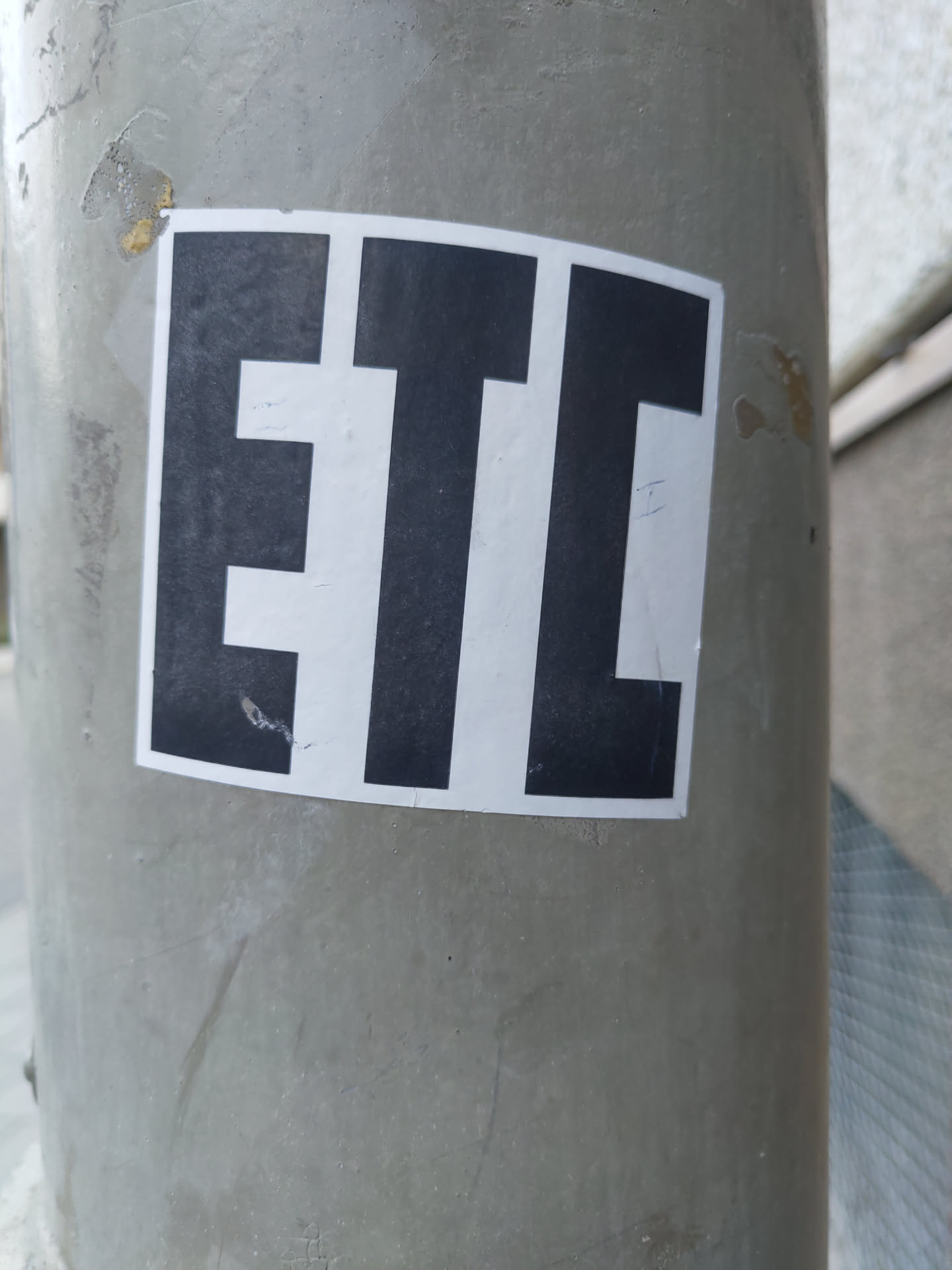 A simple sticker with the letters ETC in black on a white background. The sticker is attached to a grey pole.