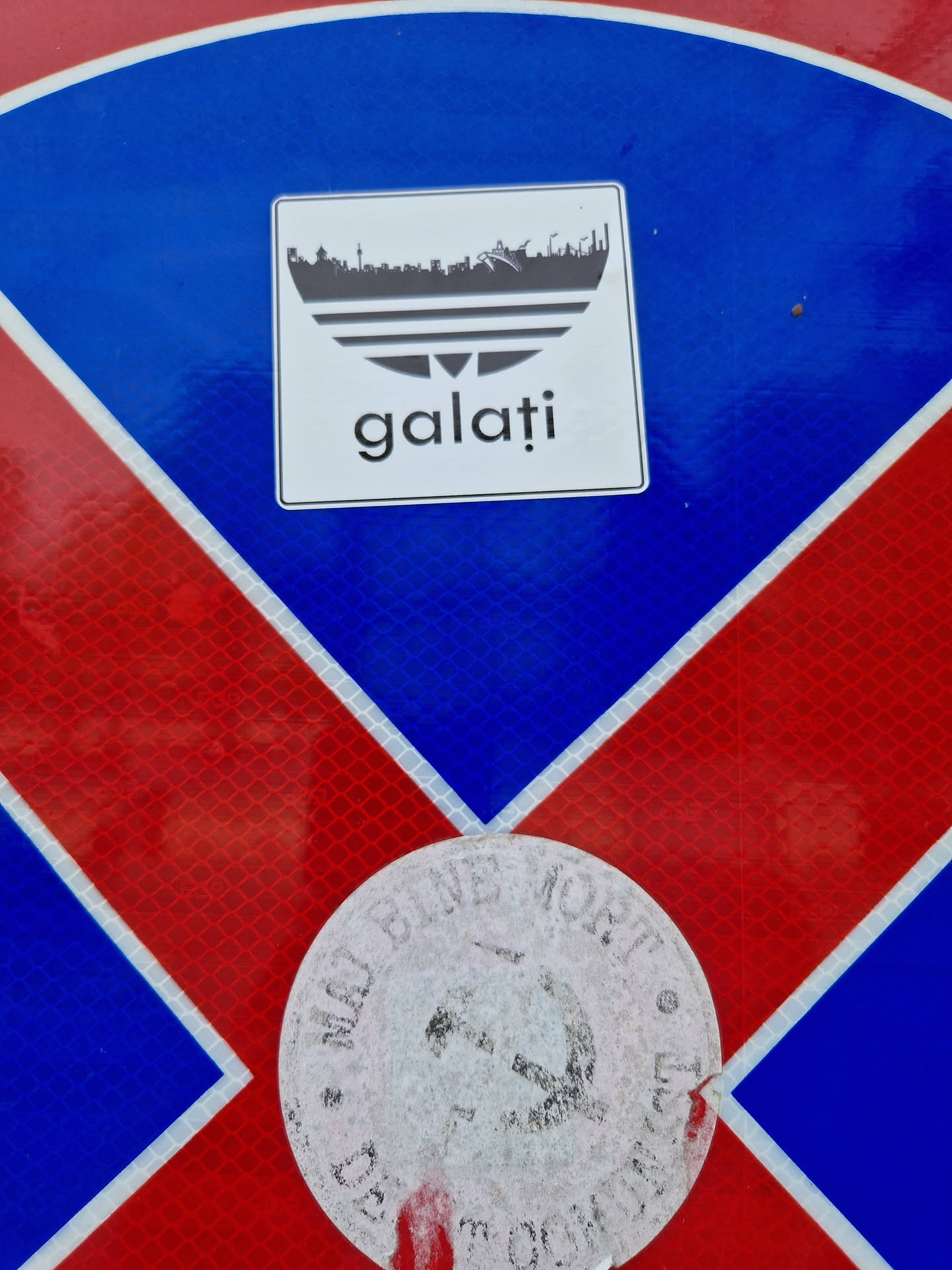 A sticker featuring a city skyline incorporated into an Adidas logo, with the word 'galati' written below. It is placed on a red and blue surface, possibly a traffic sign.