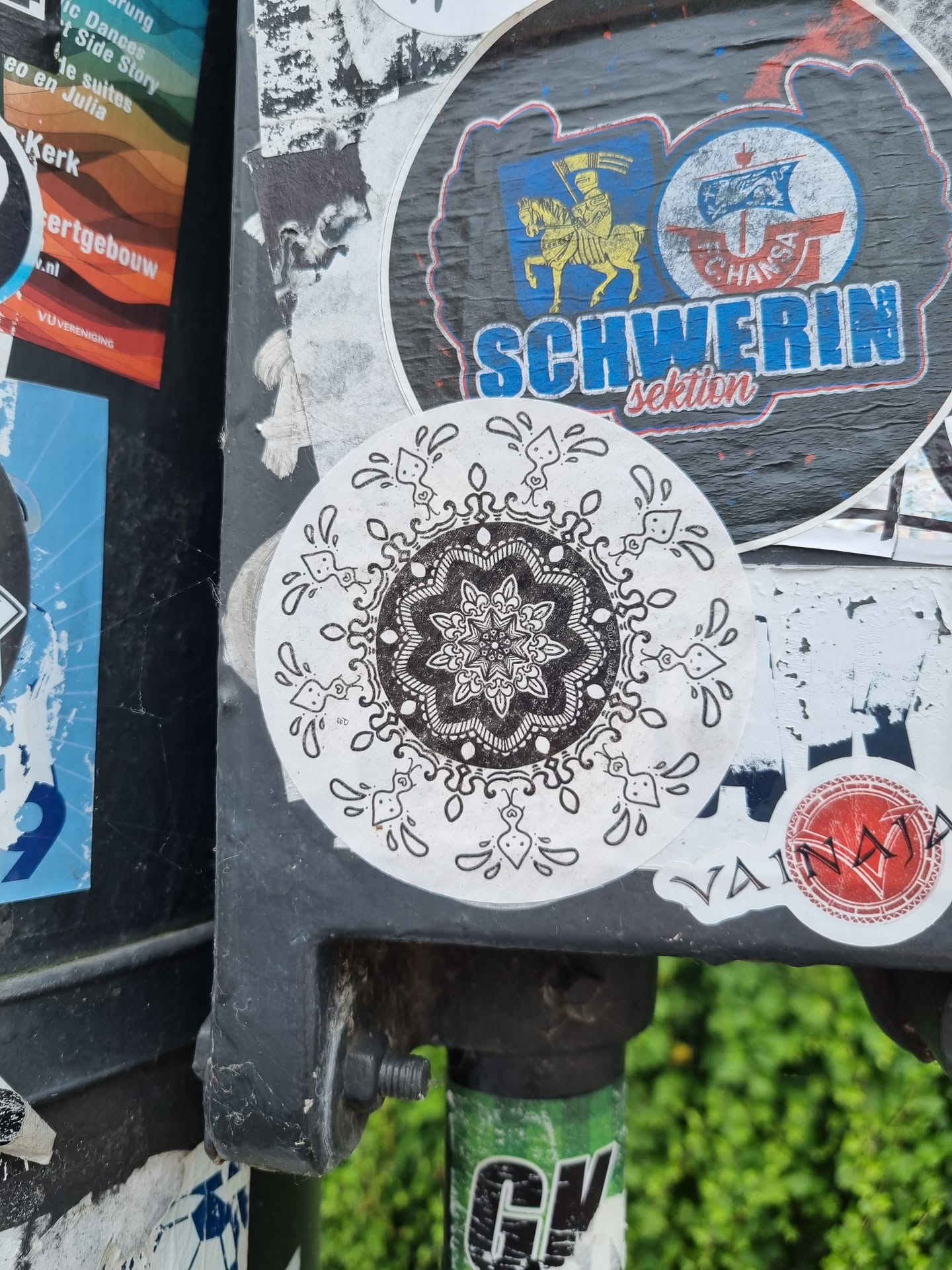 A round sticker with a detailed mandala design in black and white. The mandala is surrounded by a border of stylized droplets. The sticker is affixed to a dark gray pole, which also has other stickers on it.