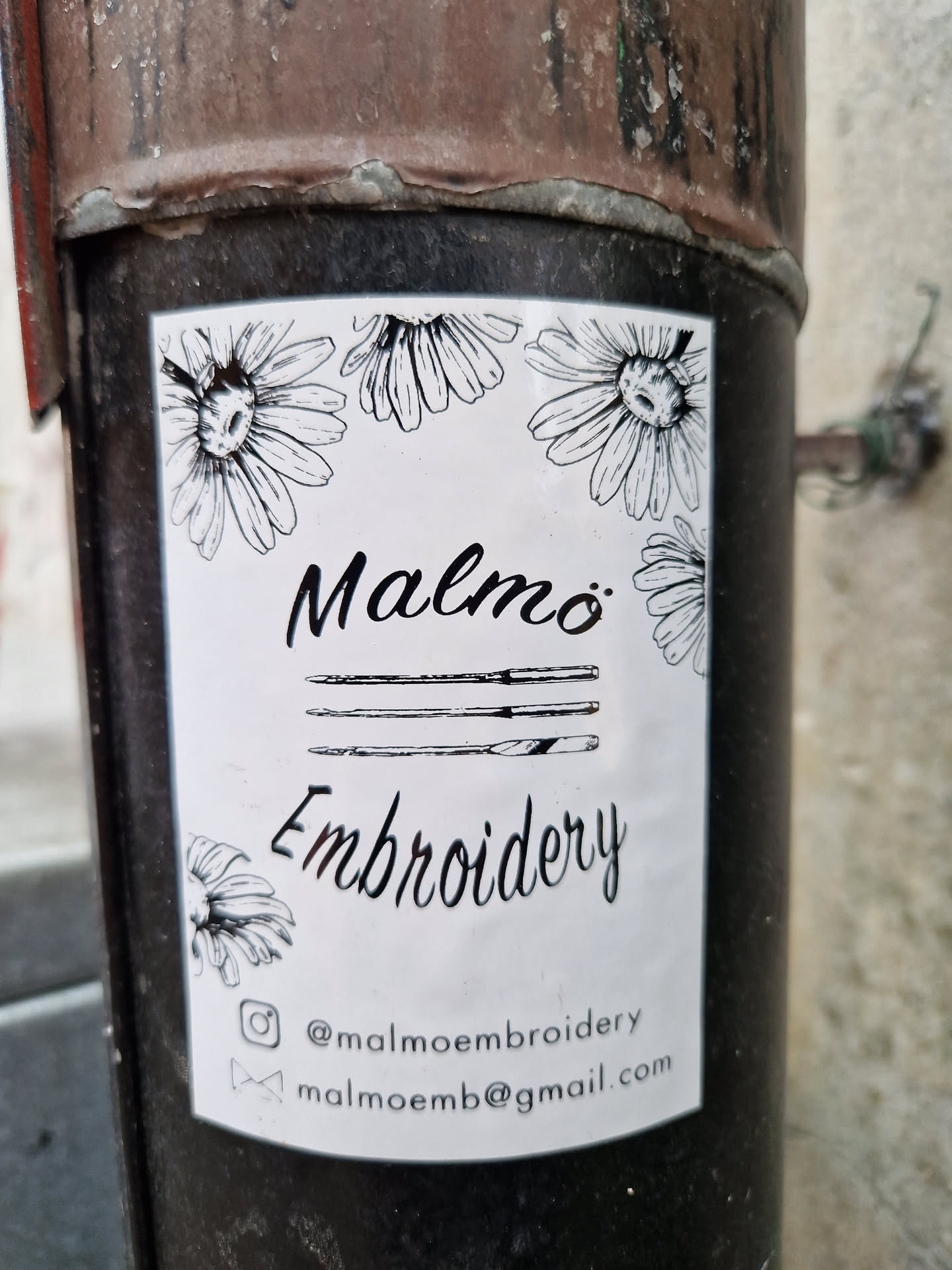 A white sticker with black ink drawings of daisies and embroidery needles.  The text on the sticker says "Malmö Embroidery" with a social media handle and email address.
