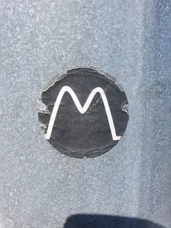 A round, black sticker with a white 'M' in the center. The sticker shows some signs of wear and tear, with edges peeling up from the surface.  It's affixed to a textured, gray metallic surface.