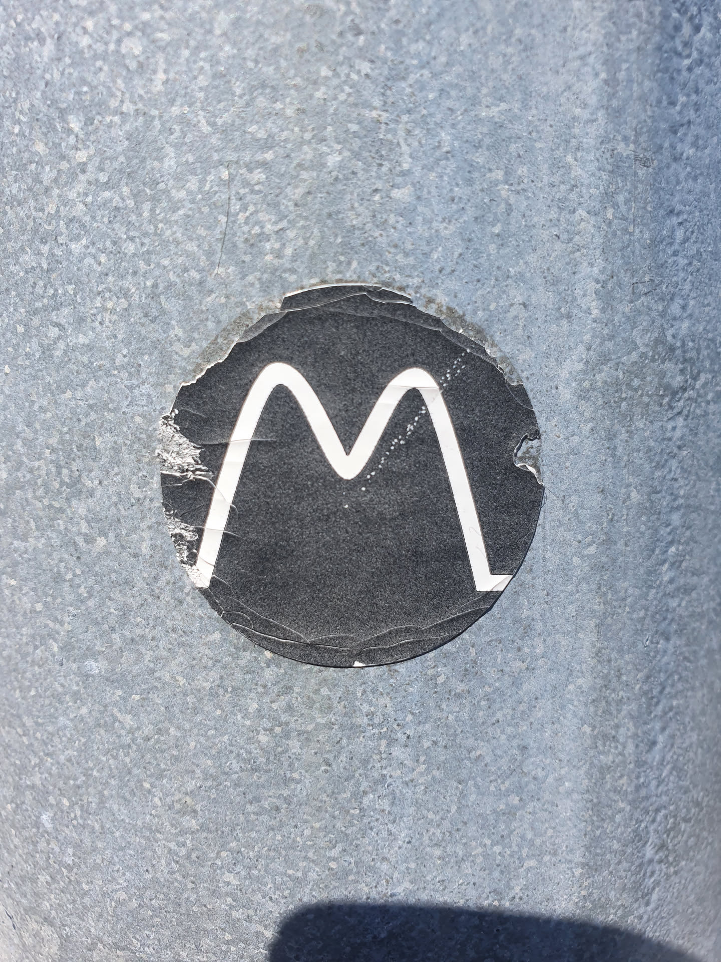 A round, black sticker with a white 'M' in the center. The sticker shows some signs of wear and tear, with edges peeling up from the surface.  It's affixed to a textured, gray metallic surface.