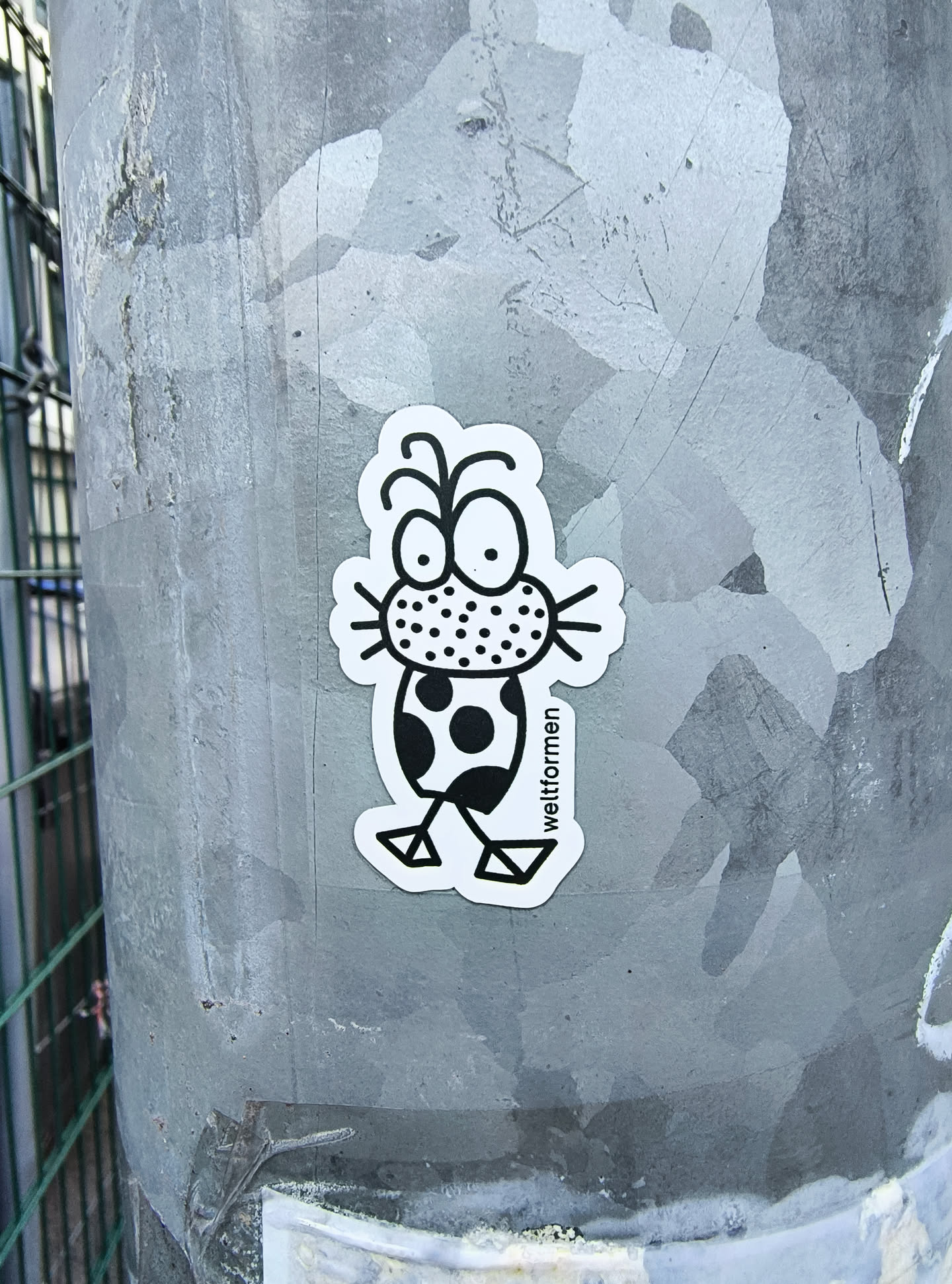 The sticker features a whimsical, abstract cartoon creature rendered in stark black and white. Its large, round eyes with prominent black pupils gaze upwards, topped by two curled antennae. The snout area is filled with a dense pattern of black dots, from which thin whiskers extend horizontally. The creature's body is rounded and adorned with irregular solid black spots, reminiscent of polka dots. Two minimalist, geometric legs, ending in triangular feet, support the body. A clean white border outlines the entire figure against the grey, textured metal pole it is adhered to. The word 'weltformen' is printed vertically along the right side of its body.