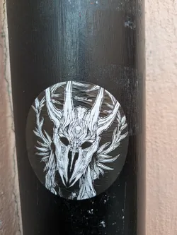This round sticker showcases a detailed illustration of a skull mask or helmet, reminiscent of ceremonial attire. The mask is surrounded by antler-like structures and foliage, creating an organic yet eerie aesthetic. The background is black, providing a stark contrast to the white linework. It's located on what appears to be a black metal or painted pole.