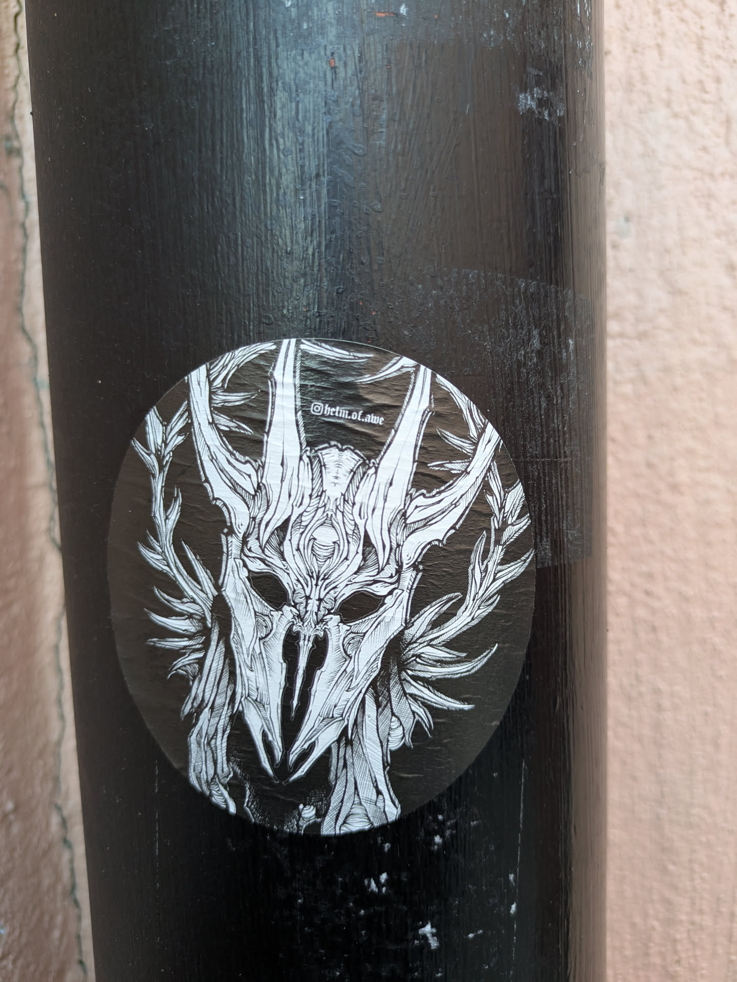 This round sticker showcases a detailed illustration of a skull mask or helmet, reminiscent of ceremonial attire. The mask is surrounded by antler-like structures and foliage, creating an organic yet eerie aesthetic. The background is black, providing a stark contrast to the white linework. It's located on what appears to be a black metal or painted pole.