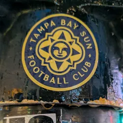 Street sticker The sticker is circular with a dark blue background and a yellow border. In the center, there is a stylized yellow sun with a face. The sun has four points extending outwards, with smaller triangular shapes within each point. The text 'TAMPA BAY SUN FOOTBALL CLUB' is written in yellow capital letters around the circumference of the sticker, separated by small yellow dots.