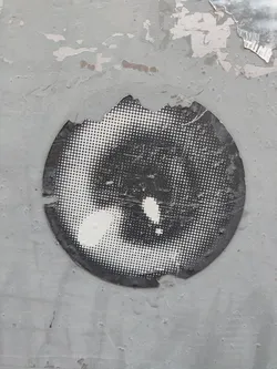 A round, black and white halftone sticker with a damaged edge. The design resembles an eye or a similar organic shape. The sticker is affixed to a weathered, gray surface.
