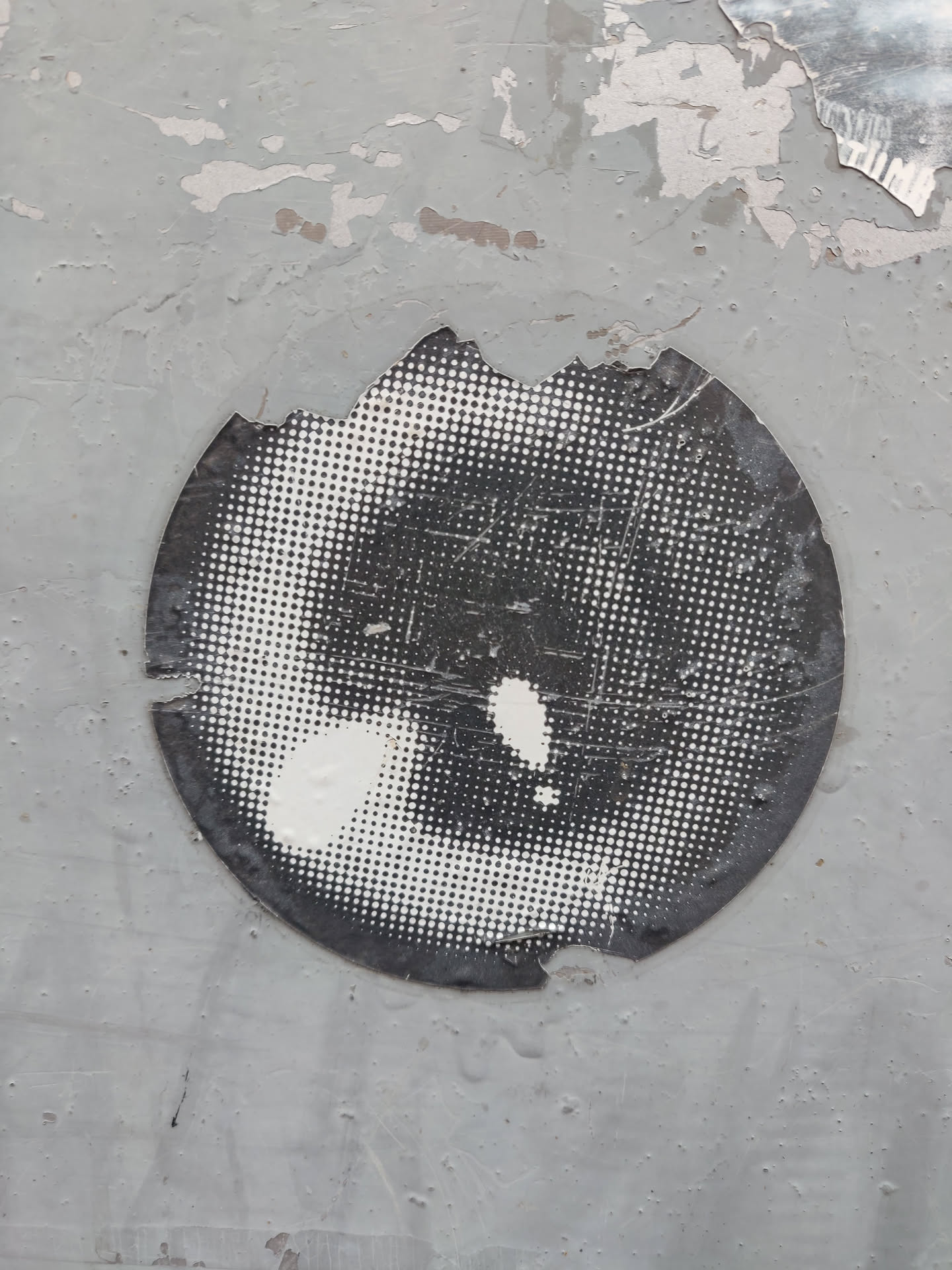 A round, black and white halftone sticker with a damaged edge. The design resembles an eye or a similar organic shape. The sticker is affixed to a weathered, gray surface.