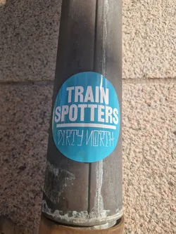 A round blue sticker with the text "Train Spotters" over "Dirty North" on a brown metal pipe.