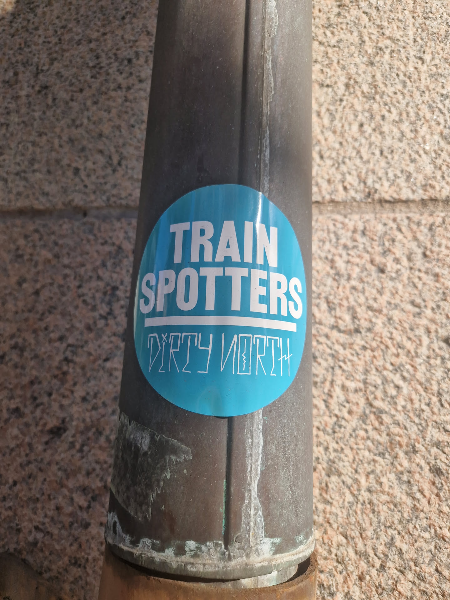 A round blue sticker with the text "Train Spotters" over "Dirty North" on a brown metal pipe.