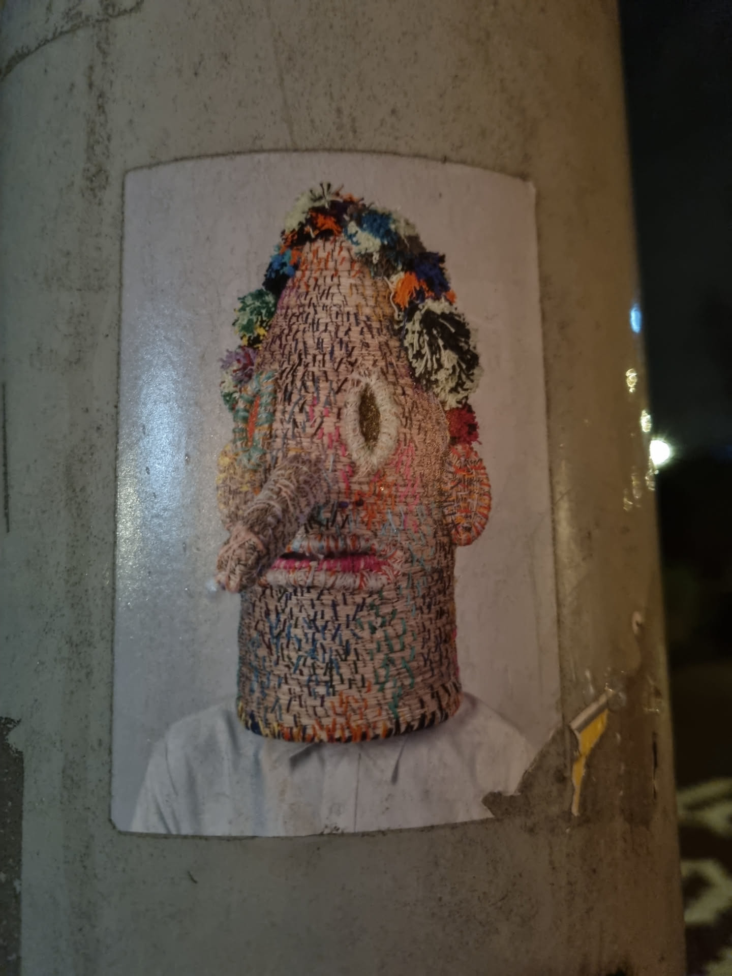 A sticker featuring a photograph of a person wearing a mask or headpiece crafted from multicolored woven textiles. The artwork displays a striking contrast between the intricate texture of the textile and the smooth background.  The face is stylized and somewhat abstract. 