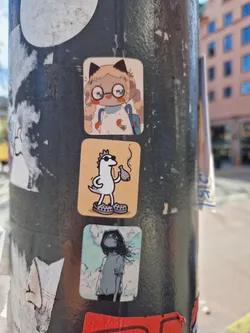 A collection of three small stickers on a lamppost. The focus sticker is a light-skinned character with blonde hair, glasses and a backpack. The sticker style is reminiscent of anime and manga. The other stickers feature a bird-like creature smoking and a character standing in the clouds. 