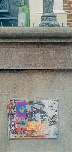 This image features a section of a textured, likely aged concrete surface, adorned with multiple layers of stickers. The primary focus is a rectangular sticker area covered with diverse images and shapes. Most notable is a purple sticker at the top left corner displaying an earth-like design. Various other stickers with geometric patterns and faded colors are scattered around it. The stickers appear faded and peeling, indicative of sun exposure and time.