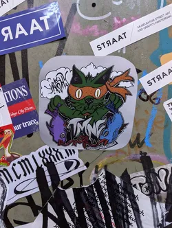 A sticker featuring an illustration of a green cat with an orange ninja mask, holding two fish. The style is cartoonish and playful. The words "TIME WARP" are subtly incorporated into the design. The background is light gray.