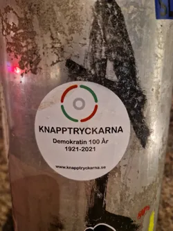 A sticker commemorating 100 years of democracy in Sweden (1921-2021).  It features a logo with green, red and white circular elements. The text on the sticker reads "KNAPPTTRYCKARNA" and "Demokratin 100 år 1921-2021", with a website address at the bottom: www.knapptryckarna.se