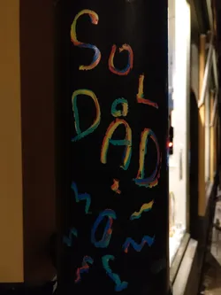 A black cylindrical sticker with the words "Sol Dad" written in colorful, childlike handwriting.  It also features some abstract, wavy lines in matching colors.