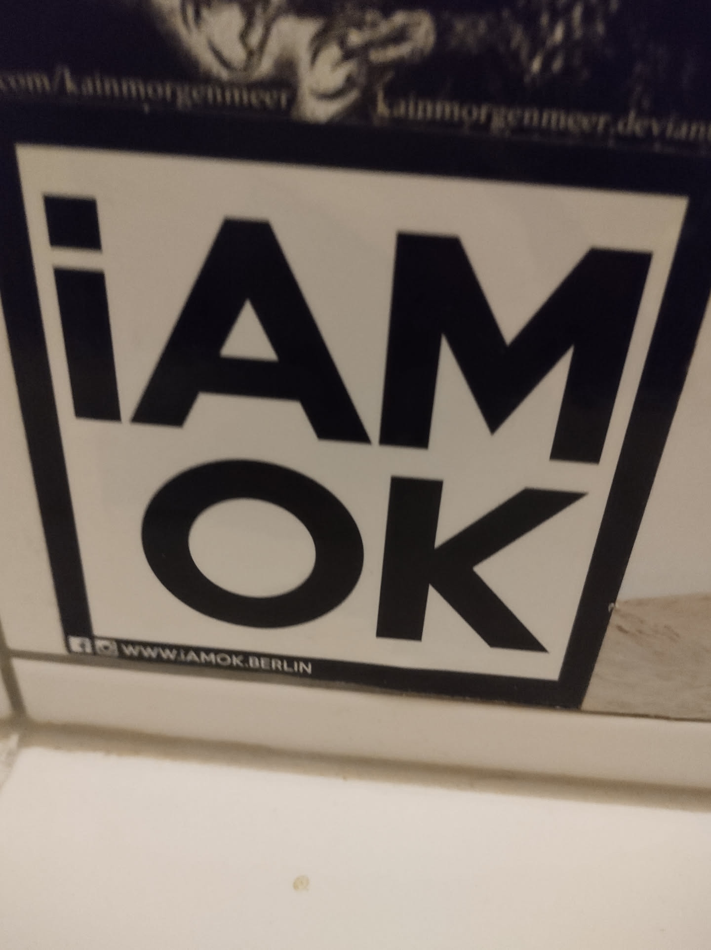 Black and white sticker with the text "i am ok" and a social media link at the bottom.