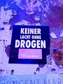 A dark sticker on a purple wall with the text: "KEINER LÄCHT OHNE DROGEN" and the bottom text: "Chill mal Berlin".