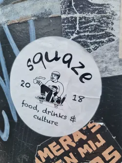 Round white sticker with black ink illustration of a person on a skateboard holding a cup. Text says "Squaeze" in a stylized font, and "20" and "18" are written on either side of the illustration. Under the image it says: "food, drinks & culture"