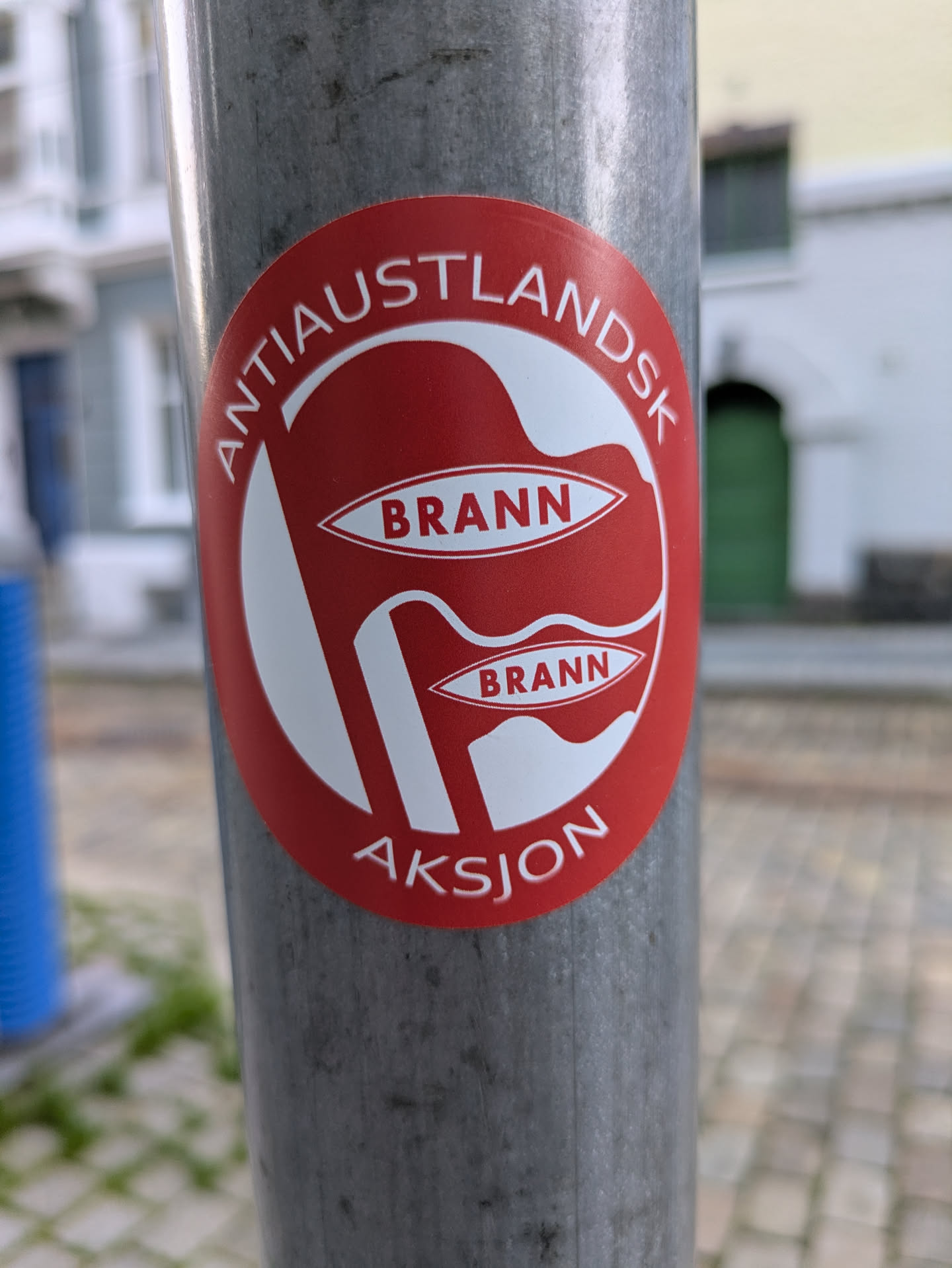 A vibrant red circular sticker with white text and graphics is affixed to a grey metal pole. The sticker features two stylized white flag or banner shapes, each containing the word "BRANN" in red text. Arched above the central design is the white text "ANTIAUSTLANDSK", and below it, also in white, is "AKSJON". This sticker appears to be related to the SK Brann football club from Bergen, Norway, and possibly an 'Anti-Eastland' regional identity or rivalry action.