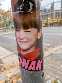 The sticker depicts a color photographic portrait of a smiling young boy with reddish-brown hair cut into bangs, cheerfully winking one eye. He is wearing a dark garment with a visible red collar. Underneath his portrait, the text 'ONAK' is printed in large, bold, uppercase pink letters. The sticker is affixed vertically to a cylindrical metal pole and shows signs of peeling and weathering along its top edge.