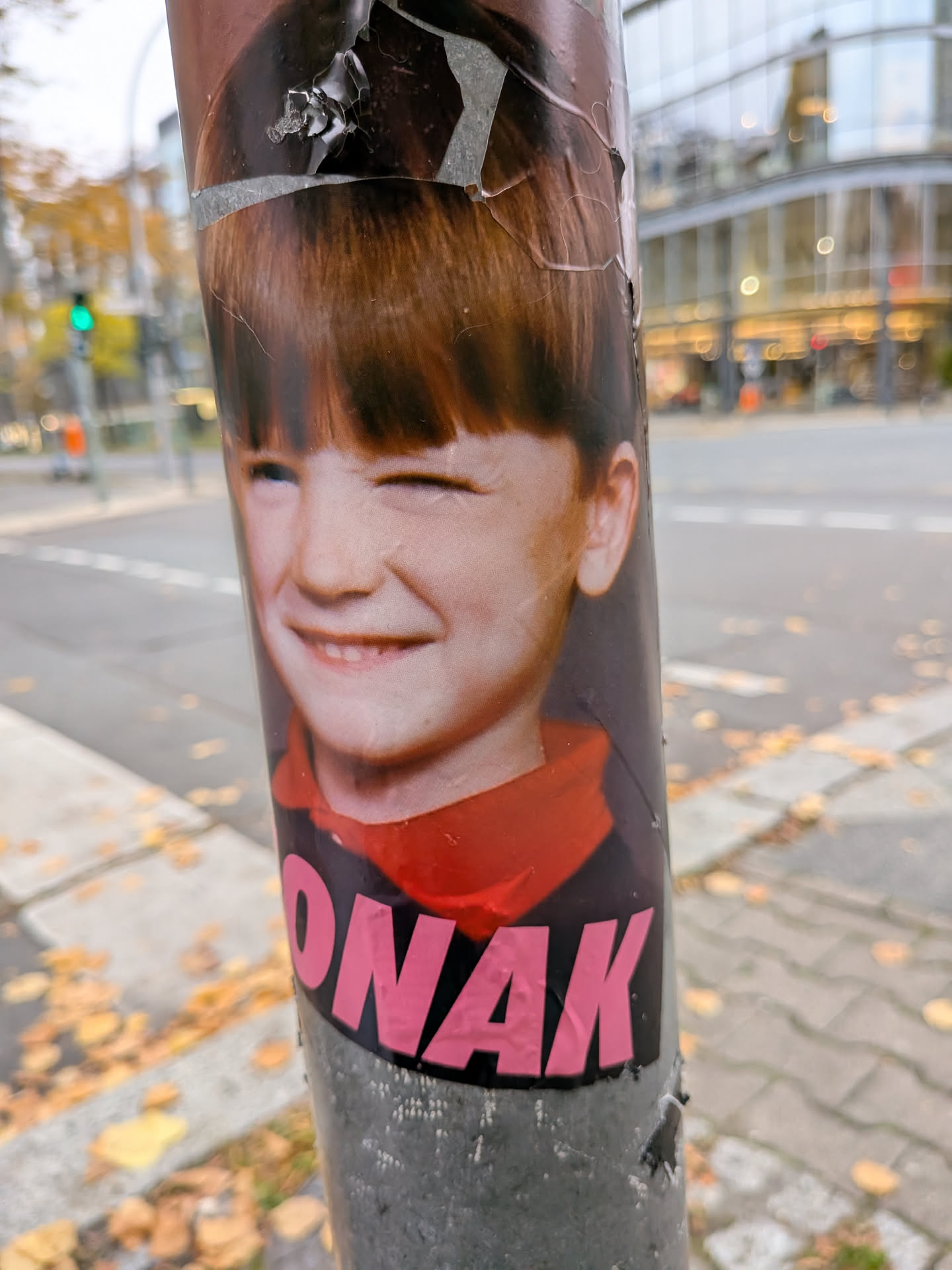 The sticker depicts a color photographic portrait of a smiling young boy with reddish-brown hair cut into bangs, cheerfully winking one eye. He is wearing a dark garment with a visible red collar. Underneath his portrait, the text 'ONAK' is printed in large, bold, uppercase pink letters. The sticker is affixed vertically to a cylindrical metal pole and shows signs of peeling and weathering along its top edge.