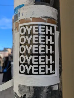 The sticker features the text 'OYEEH.' repeated four times in white letters against a black background. A small skull is next to each repetition of the text. The sticker is on a pole.