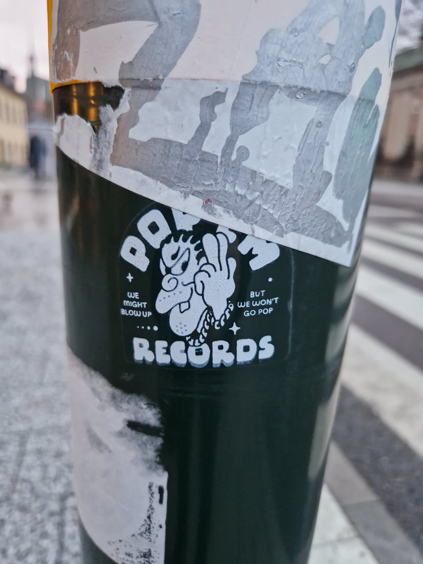 A sticker featuring the POVM Records logo, which includes a cartoon character and the text WE MIGHT BLOW UP... BUT WE WON'T GO POP. The sticker is placed on a dark green pole.
