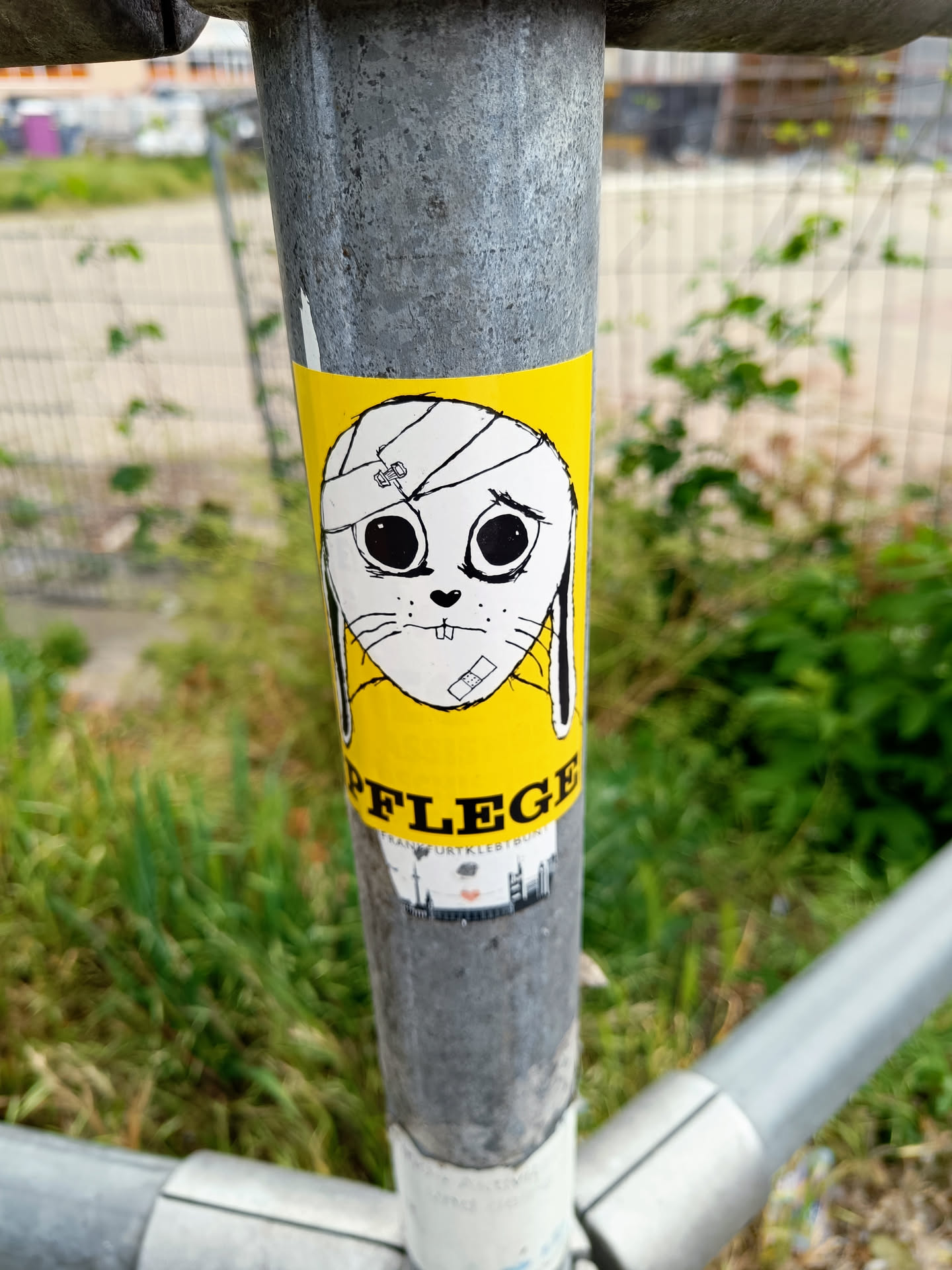 A yellow sticker featuring a cartoon bunny with a bandage on its head. The word 'PFLEGE' (care) is written below the bunny, along with a small cityscape graphic. The style is simple and slightly melancholic. The sticker is affixed to a metal pole outdoors.