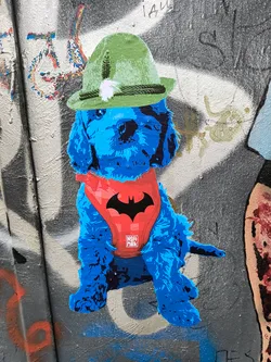 A sticker featuring a blue dog wearing a red batman harness and a green hat. The sticker is on a wall with graffiti.