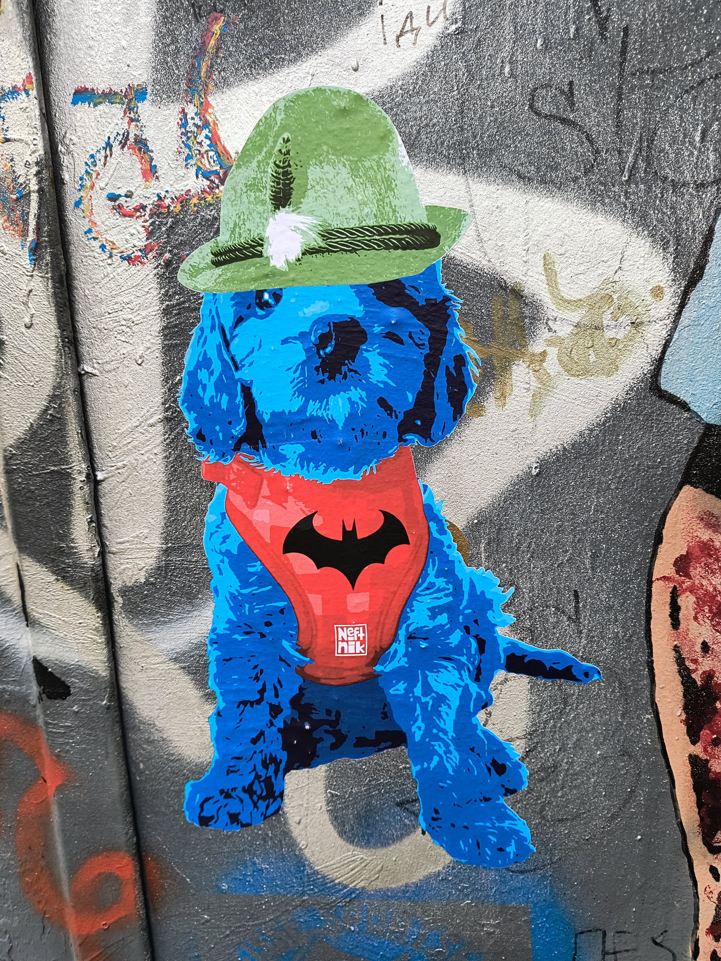 A sticker featuring a blue dog wearing a red batman harness and a green hat. The sticker is on a wall with graffiti.
