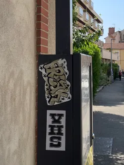 The image features two distinct stickers applied to a dark, possibly metallic, utility box or pole. The top sticker is larger and irregularly shaped, cut around a graffiti-style word. The word, which appears to be 'PREST', is rendered in a bubbly, stylized font with a light brown or beige fill, outlined in black. Small black stars are scattered around and within the letters, all set against a white background. The sticker shows some signs of wear, with slight peeling at the edges. Below it, a rectangular white sticker displays the letters 'VHS' in a bold, blocky, black font with a white outline, evoking a retro aesthetic.