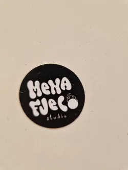 A black and white circular sticker with the text "Mena Fuego Studio" in a stylized font.  A small flame design is incorporated into the logo.