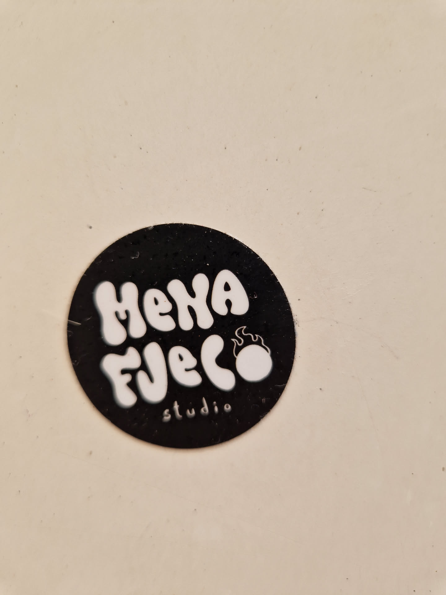 A black and white circular sticker with the text "Mena Fuego Studio" in a stylized font.  A small flame design is incorporated into the logo.