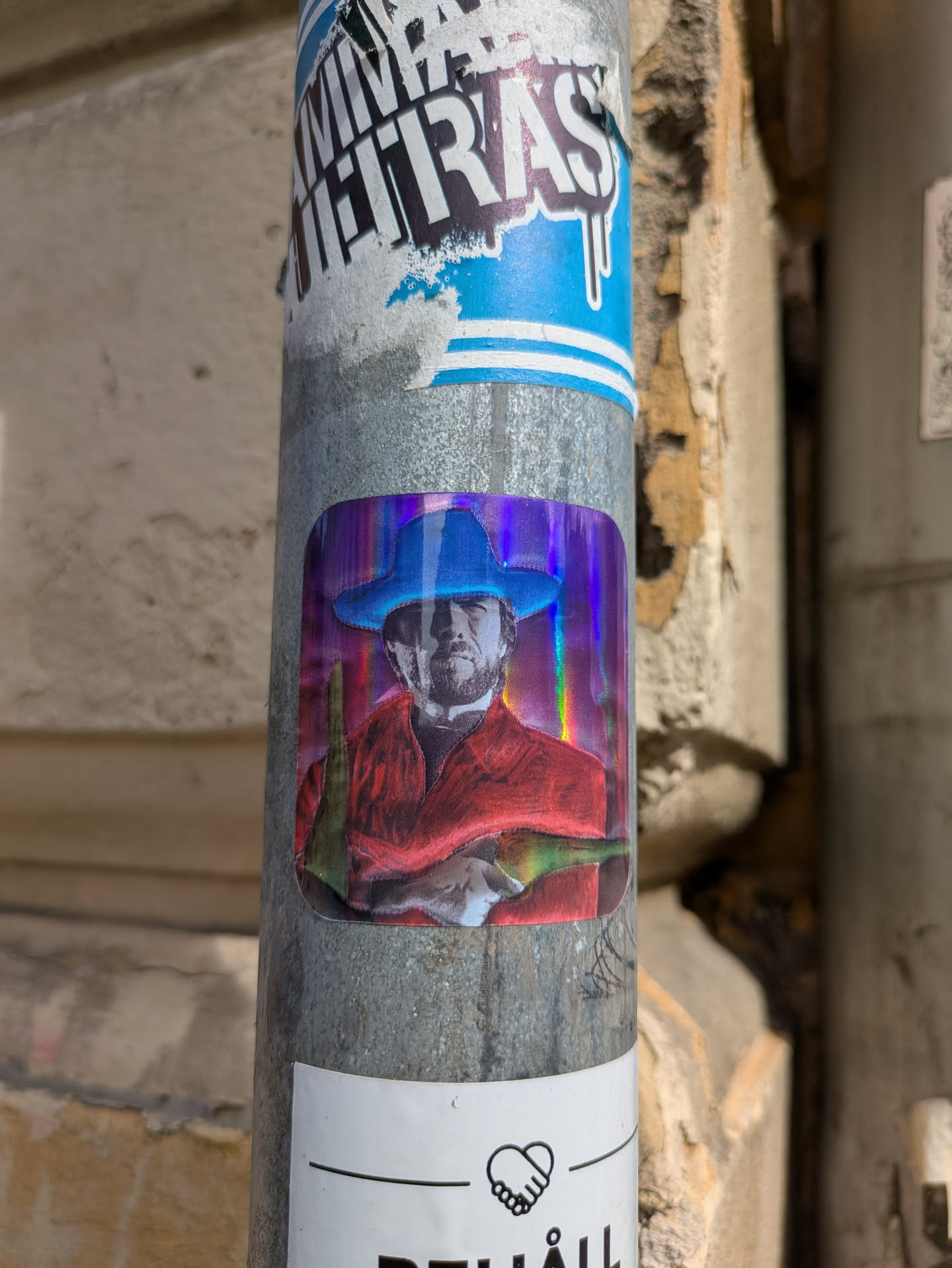 A holographic sticker adhered to a metal pole shows a man in a cowboy hat holding two bottles of wine. The sticker is holographic, giving it a shiny, rainbow effect. The man is wearing a red shirt.