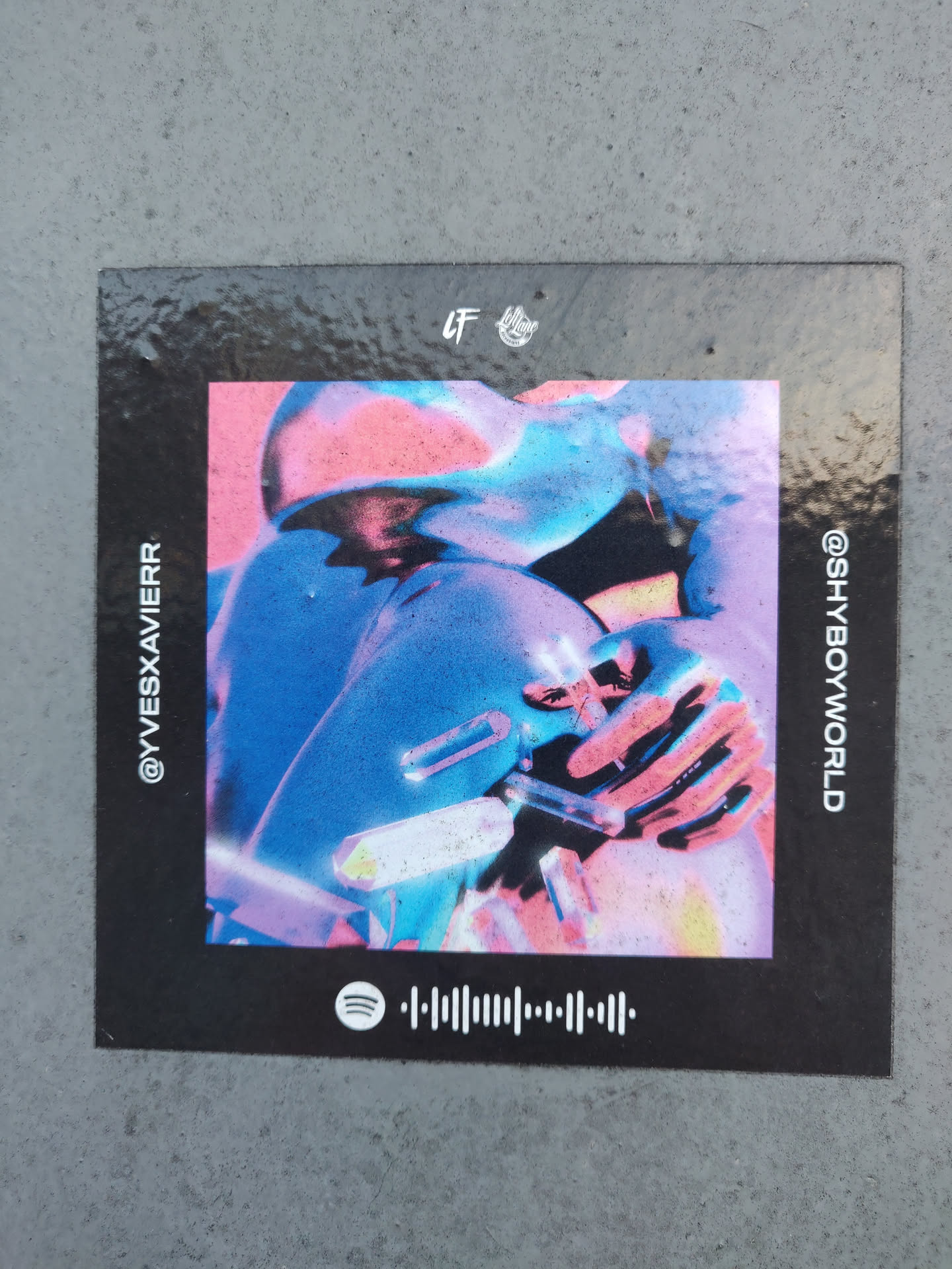 A black sticker featuring a surreal abstract artwork with pink, blue and purple tones. It has the artists' social media handles @yvesxavier and @shboyworld printed on the left and right sides respectively. A Spotify code is also present at the bottom.  The sticker appears to be glossy.