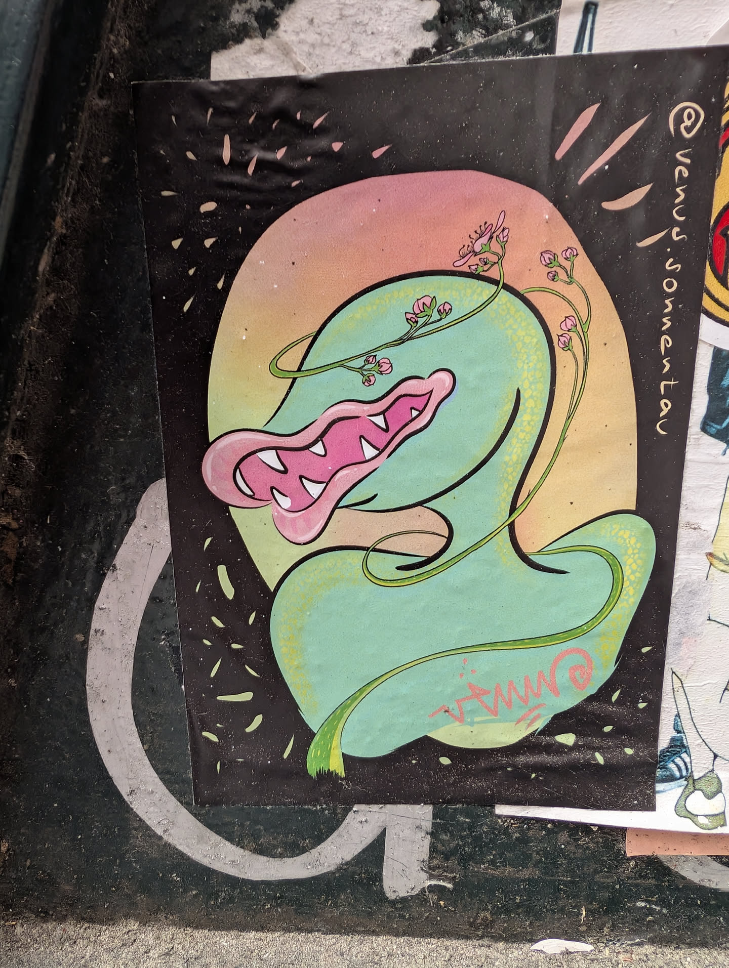 A colorful sticker features a whimsical creature with a green, elongated body and a pink mouth with sharp teeth. Flowers and vines intertwine around the creature. The background is a mix of black with scattered white dashes and a soft gradient of pink and orange. The sticker is adhered to a surface, possibly a street fixture, with other stickers partially visible.