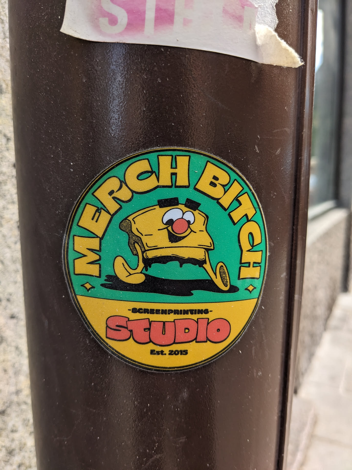 A round sticker featuring a cartoon illustration of a piece of toasted bread with a face. The sticker is predominantly teal green with yellow and orange accents. Text on the sticker reads: 'MERCH BITCH' in a circular design and 'SCREENPRINTING STUDIO Est. 2015' in smaller text below the character. The sticker is affixed to a brown pole.