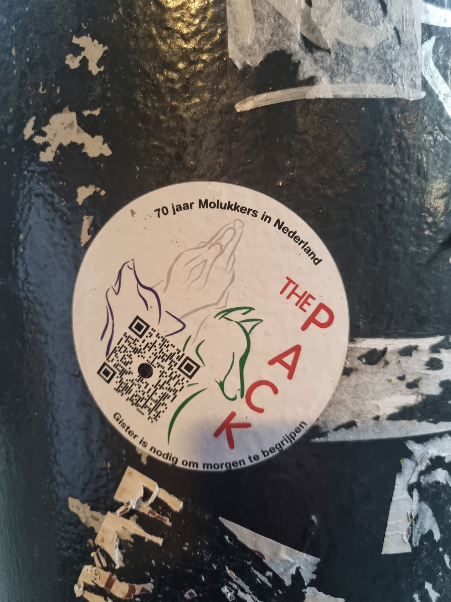 Round sticker commemorating 70 years of Moluccans in the Netherlands. It features a QR code, praying hands, and the text "The Pack" in a stylized way.  The bottom text translates from Dutch to "Yesterday is necessary to understand tomorrow."
