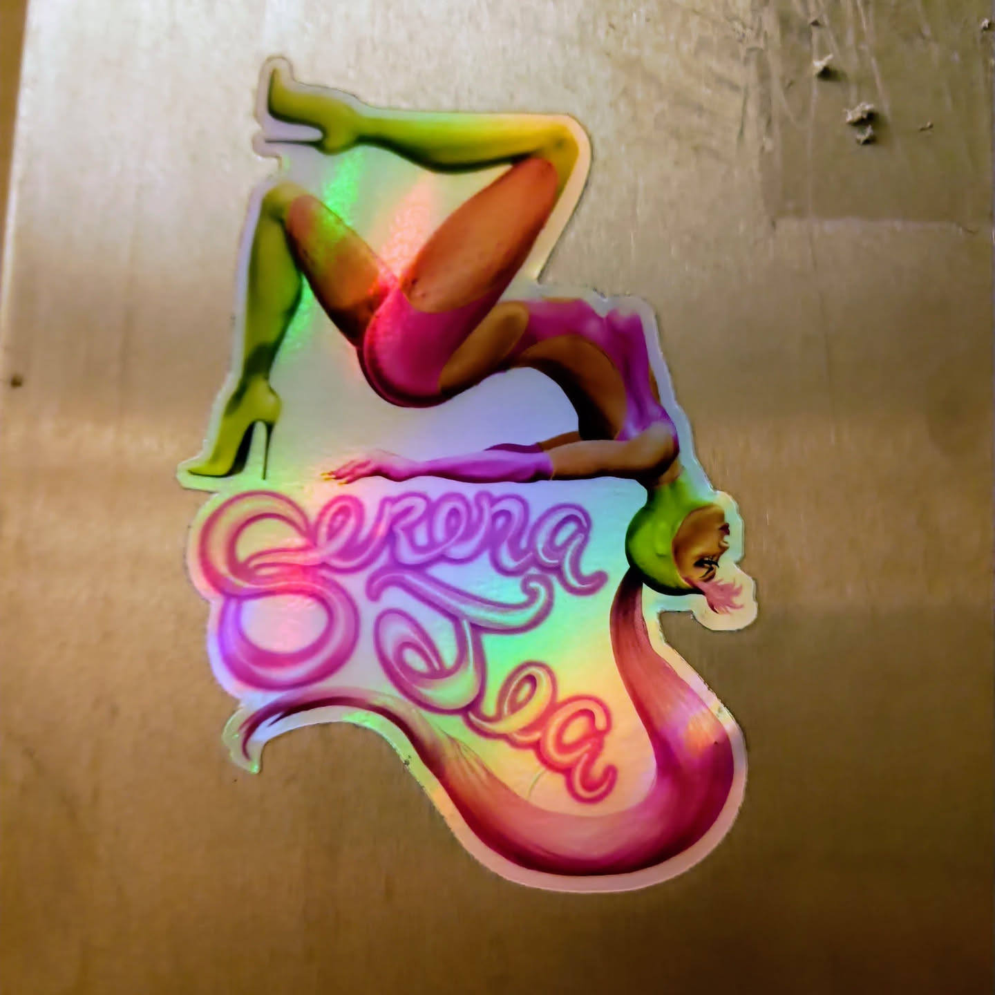Holographic sticker featuring an illustration of Serena DeSea in a dynamic pose. The sticker has a vibrant iridescent finish, showcasing a spectrum of colors.  Her name is written in a stylized font below her.