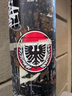 The sticker is circular, featuring a prominent black double-headed eagle with white outlines, centered within a white shield-like shape. This shield is encircled by a detailed white laurel wreath. The entire design is set against a vibrant red background, which is bordered by a thin white line and then a thin black outer ring. The sticker is partially covered by a strip of clear tape across its upper section, adhering it to the rusty black pole.