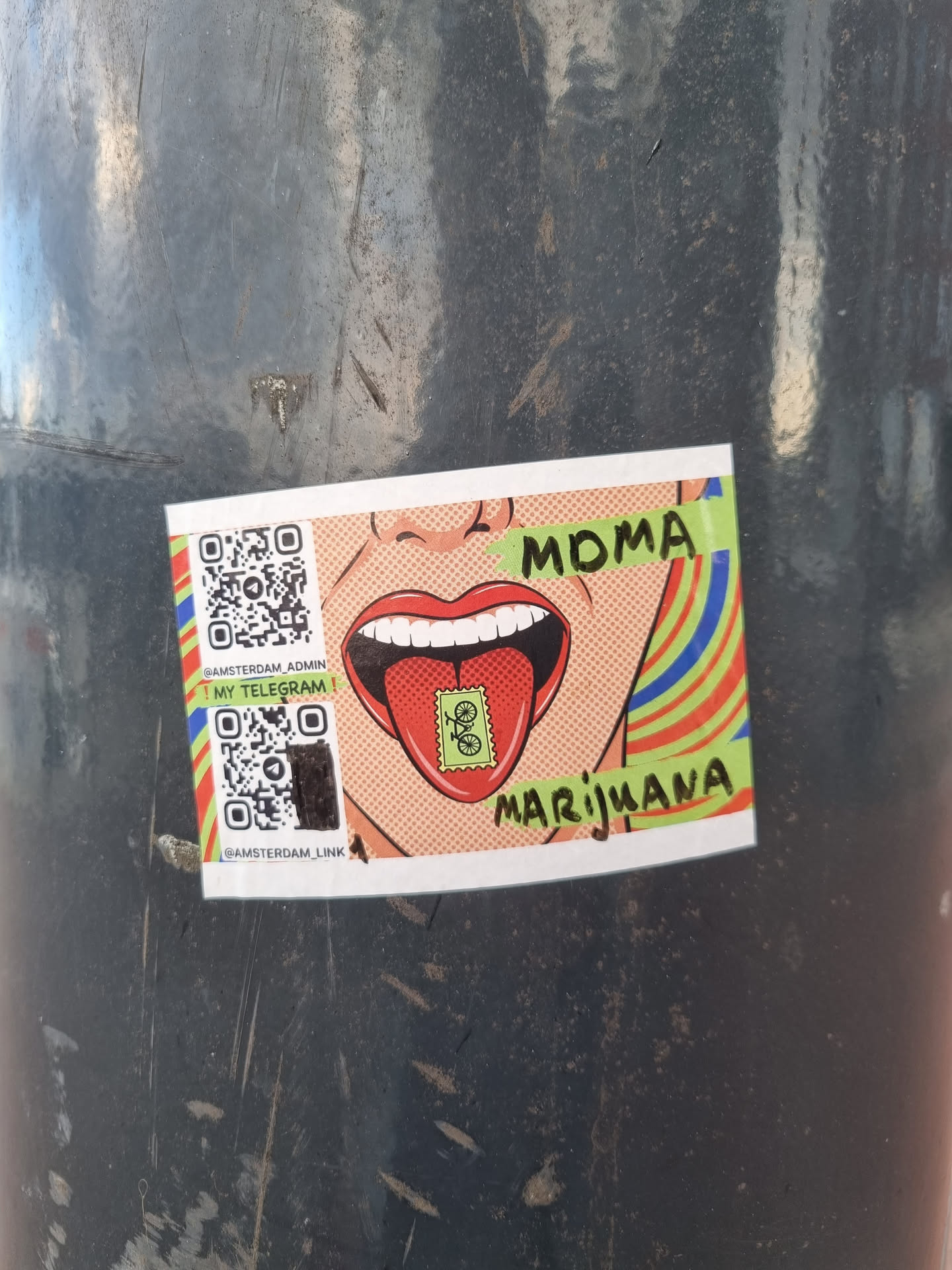 A sticker featuring a pop art style illustration of a woman's mouth with a tongue sticking out, holding a stamp with a bicycle on it. The text "MOMA" and "Marijuana" is visible on the sticker.  Two QR codes are also included, linking to a Telegram channel.