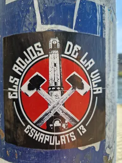 A sticker with a red circle and crossed axes in the center. There is a tower on the axes. The text around the circle says Els rojos de la Vila Eskapulats 13.