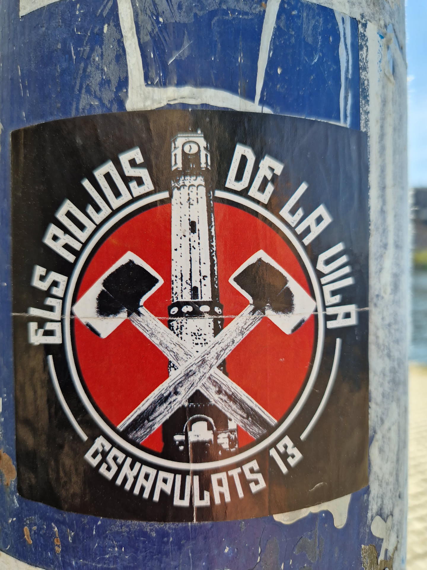 A sticker with a red circle and crossed axes in the center. There is a tower on the axes. The text around the circle says Els rojos de la Vila Eskapulats 13.