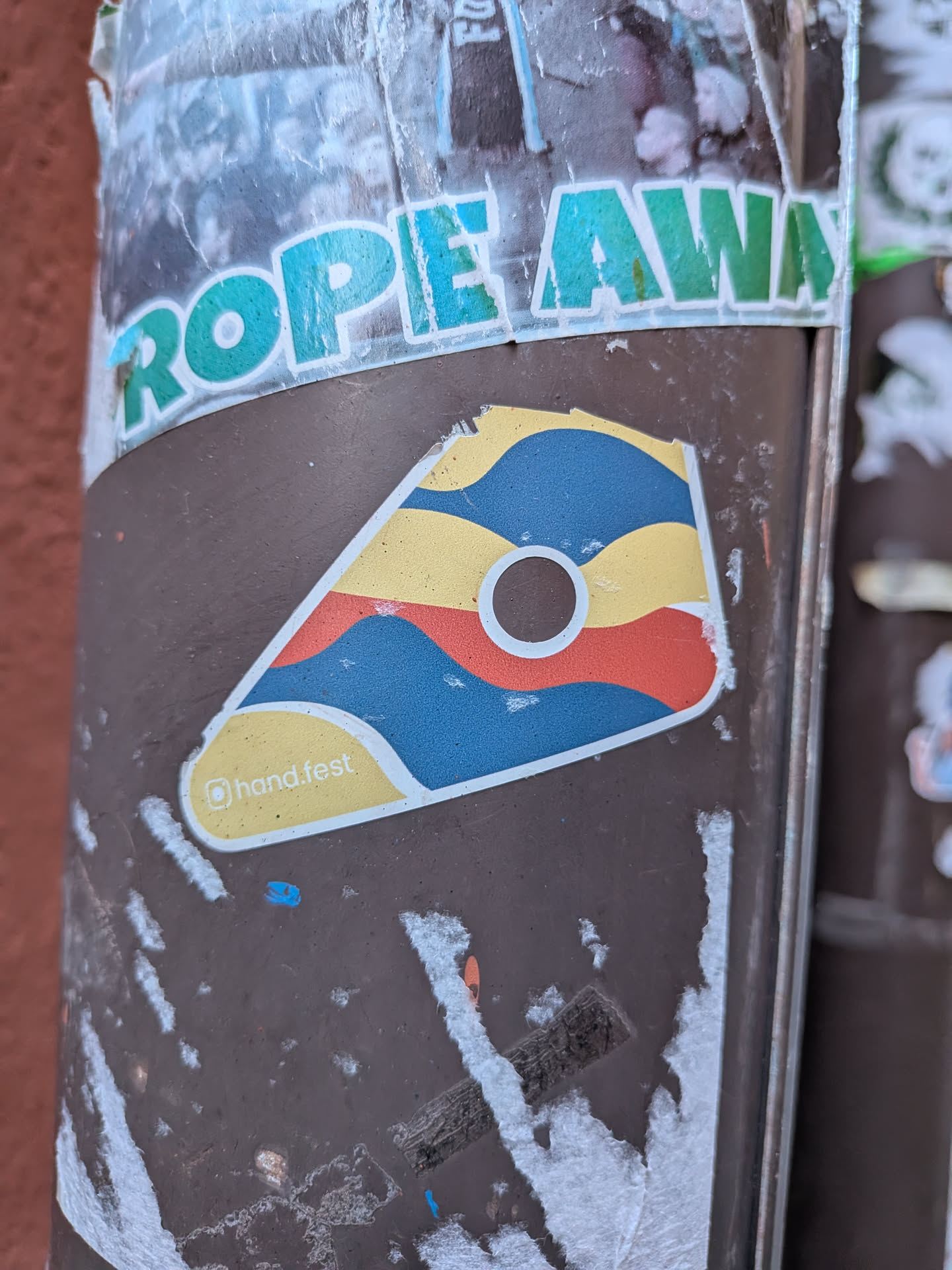 The sticker is an irregular, somewhat triangular or trapezoidal shape with rounded corners, resembling a stylized guitar pick or shield. It features a bold, abstract design composed of wavy, horizontal bands of color: yellow at the top, followed by blue, then red, and another blue band at the bottom. A prominent white circle with a dark brown center is located in the upper-middle section of the sticker. The entire design is outlined in white. In the bottom-left corner, within a yellow section, is the text '@hand.fest'. The colors are bright and distinct, giving it a modern, graphic appearance.