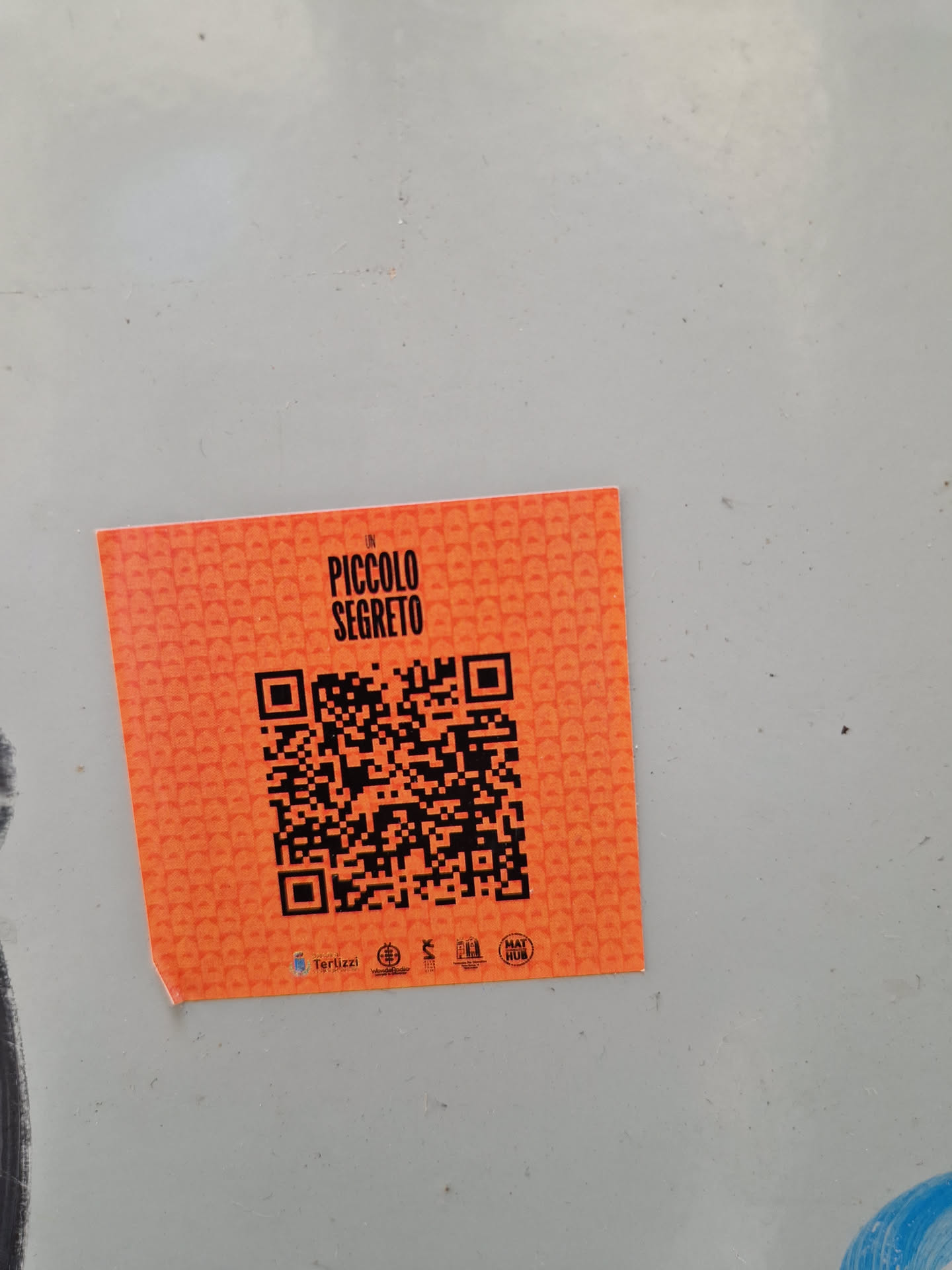 Orange square sticker with a QR code in the center. The text "Un Piccolo Segreto" is printed on top of the QR code. There are some small logos at the bottom of the sticker.