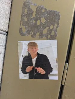 The sticker features a photo of a fair-skinned person with short blond hair wearing a dark jacket and white shirt. The background appears to be a modern interior with white walls and geometric elements. The sticker is adhered to a somewhat worn metal surface.