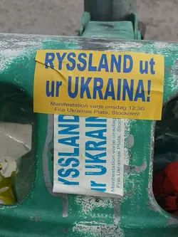A rectangular yellow sticker with bold blue text, reading 'RYSSLAND ut ur UKRAINA!' (Russia out of Ukraine!). Below this, smaller black text details a weekly demonstration: 'Manifestation varje onsdag 12.30 Fria Ukrainas Plats, Stockholm!' (Demonstration every Wednesday 12.30 Free Ukraine's Square, Stockholm!). The sticker shows signs of wear, including a crease on the top left corner, and is affixed to a textured, chipped green surface. A similar, partially obscured sticker is visible beneath it.