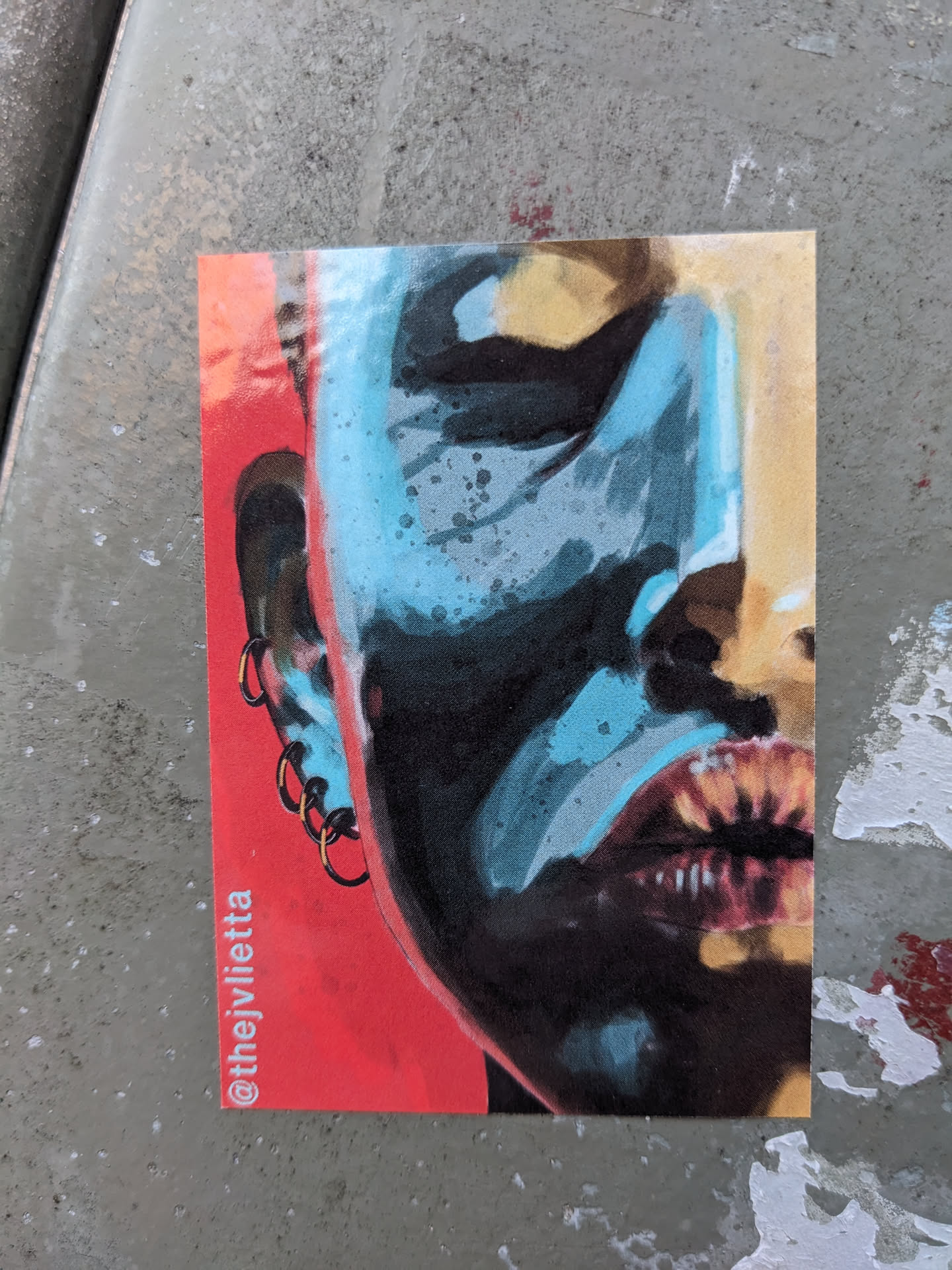 Close-up view of a sticker featuring a stylized portrait. The artwork uses a blend of teal, black, and red, creating a striking contrast. The subject's face is partially visible, with an emphasis on eyes and lips. The artist's Instagram handle is visible on the sticker's bottom left corner.